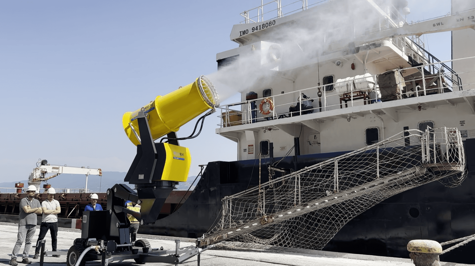 A large yellow fog cannon spraying mist over a stockpile of iron ore at a port.